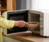 Hand placing bowl of vegetables inside microwave on wooden kitchen benchtop